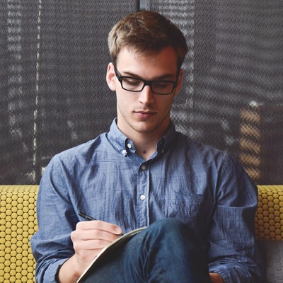 Picture Of A Young Man Wearing Glasses And Writing Notes In A Notebook