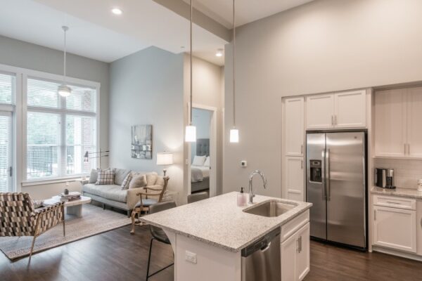 Different Angle Of The Living Room And Kitchen Area In A Harpeth Square Apartment, It Has A Sofa, Two Chairs, A Sink, A Fridge, Cabinets, Grey Walls And Windows Looking To The Balcony.