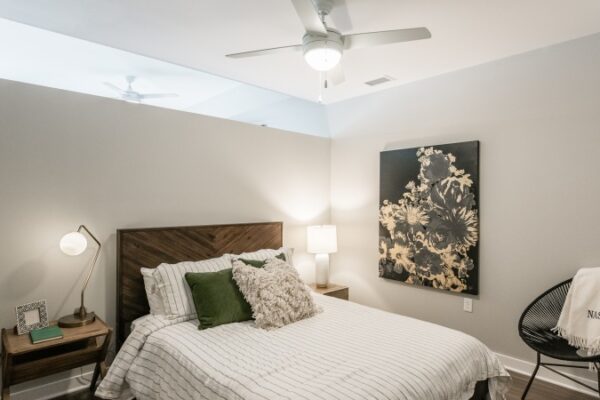 A Bedroom In A Harpeth Square Apartment. It Has A White Bed With Green And Grey Pillows, A Lamp, A Painting On The Wall, A Chair With A Blanket On It That Says Nashville.