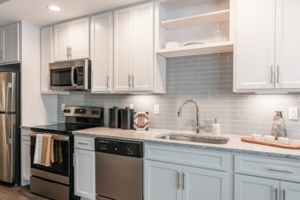 Kitchen In A Harpeth Square Apartment. It Has White Cabinets, A Metallic Fridge, Dishwasher, Electric Stove, Microwave, And Other Amenities. It Looks Ample.