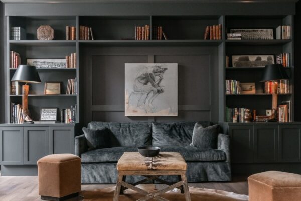 Different Photo Of The Studio Or Living Area In Harpeth Square. It Has A Large Dark Grey Sofa, Two Chairs, A Wooden Shelf With Books. The Walls Are Dark Grey.