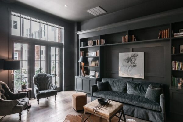 Studio Or Living Area In Harpeth Square. It Has A Large Dark Grey Sofa, Two Chairs, A Wooden Shelf With Books. The Walls Are Dark Grey And Has A Window Looking Outside.
