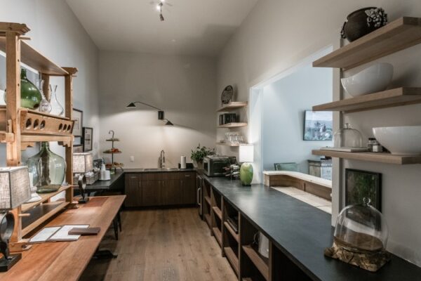 Kitchen In A Harpeth Square Apartment. It Has A Rustic, Comfy Feel. Shelves Are Made Out Of Wood. It Has Glasses, Plates, A Microwave. It Has A Sink And A Space To Look To The Living Room.