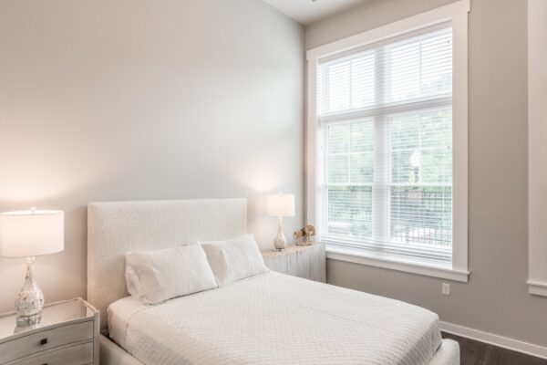 Bedroom In A Harpeth Square Apartment. It Has A Large, White Bed, Two Lamps, A Bureau, And Two Windows Looking To The Outside.