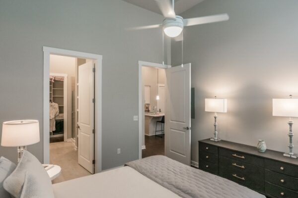 Bedroom In A Harpeth Square Apartment. It Has A Large, White Bed, Two Lamps, A Bureau, And Has Two Open Doors, One Looking At The Kitchen And The Other One To The Bathroom And Closet Area.