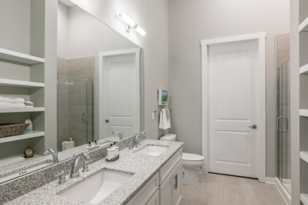 Bathroom In A Harpeth Square Bathroom. It Has A Large Mirror, Two Sinks, Towels, A A Space For Folding Clothes. It Is White And Bright.